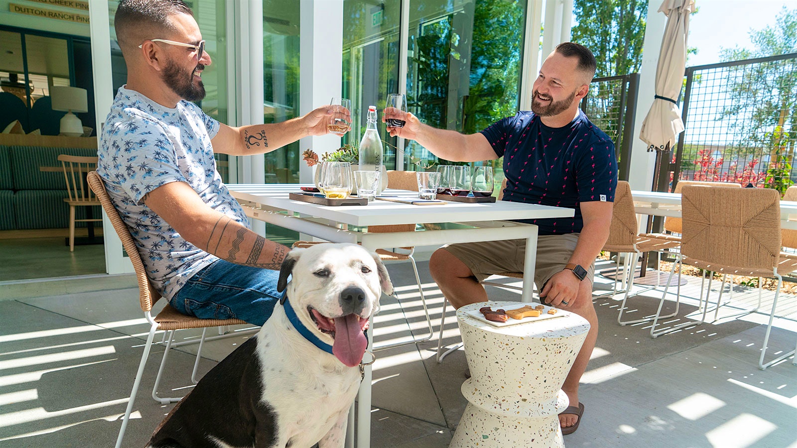 Two men toast with glasses of wine at Migration’s outdoor courtyard while a dog looks on.